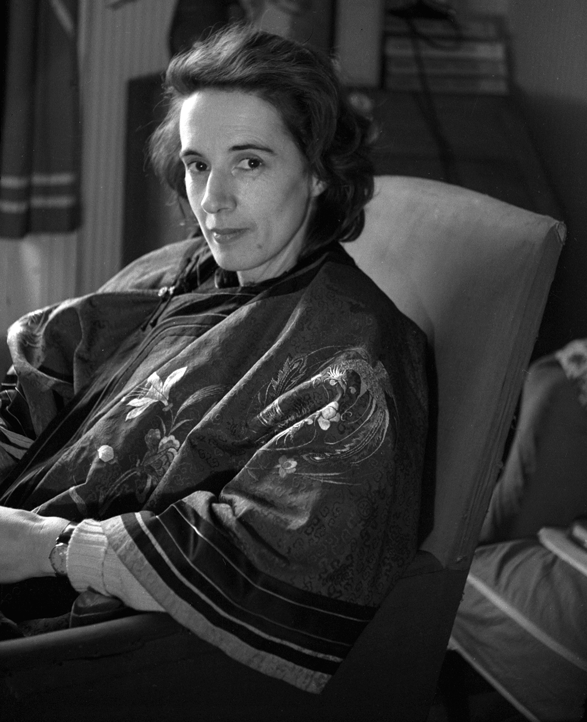 Black and white photograph of a woman sat in a chair looking out at the camera. She has shoulder length hair and wears a floral patterned jacket.